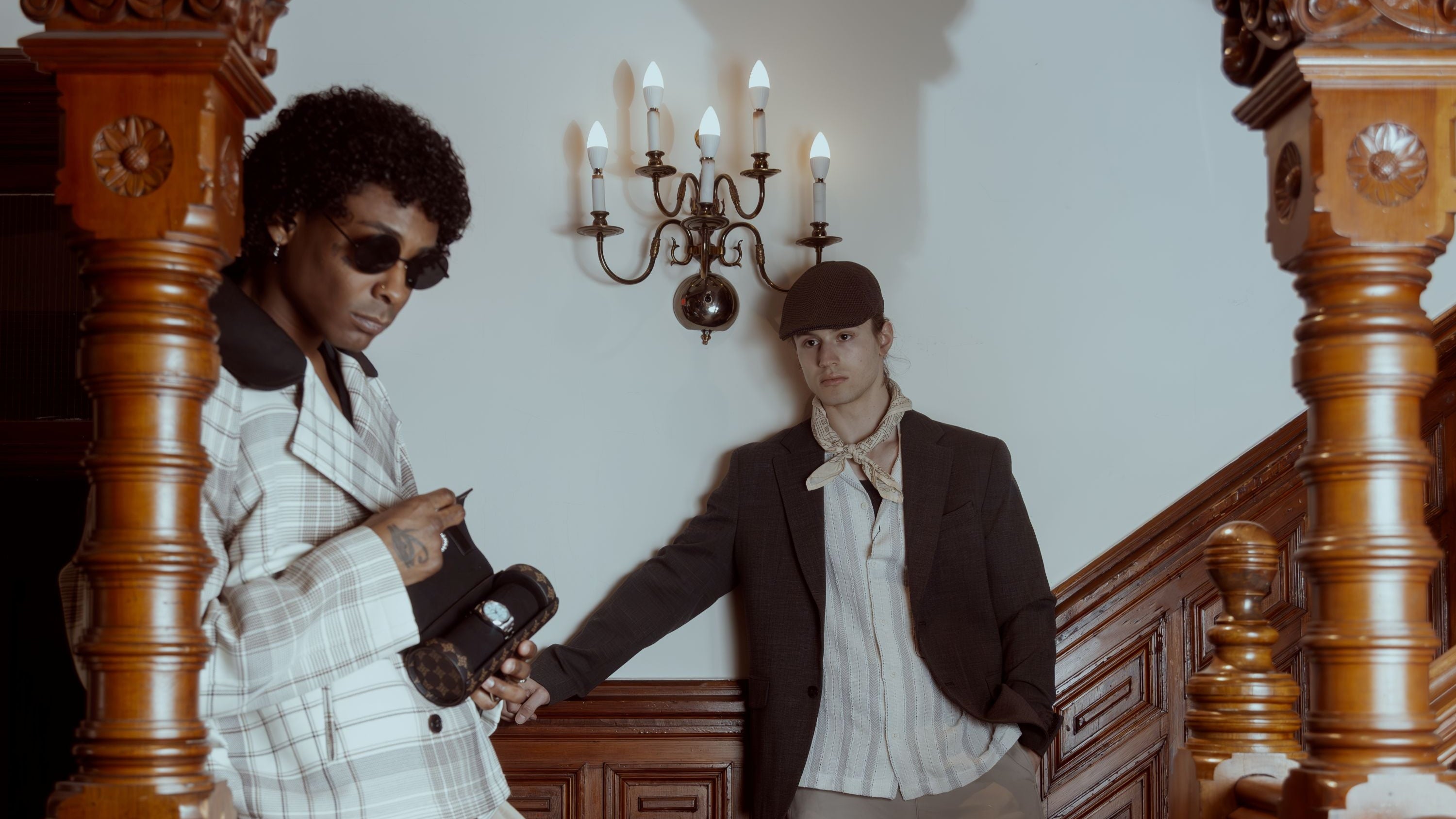 Two men standing in a grand hallway with ornate wooden railings holding a watch case